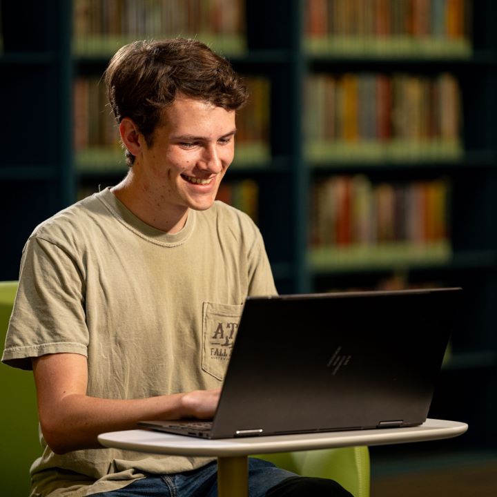 Student sits in a green chair in the Salmon Library smiling at his computer screen in front of him