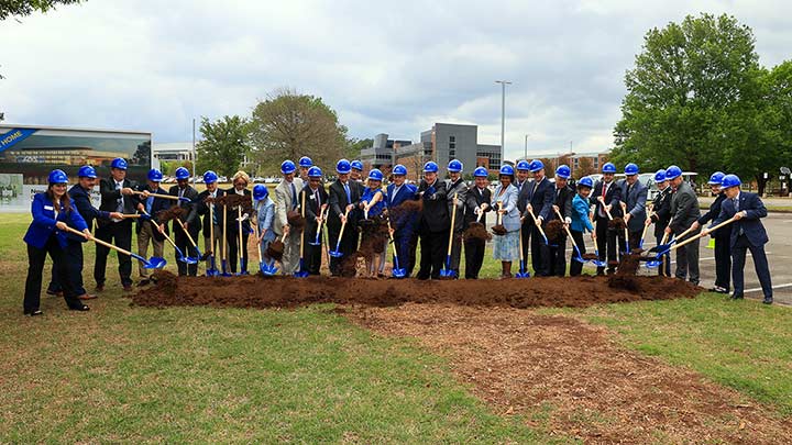 a large group of people in blue safety hats hold shovels near a pile of dirt