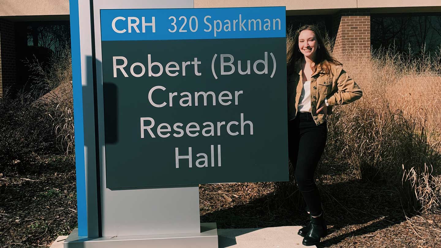 Emily Wisinski standing next to the Robert Cramer Research Hall building sign.
