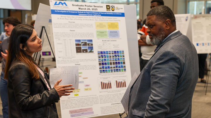 A graduate student discusses their research at a poster session, featuring various charts and visuals about AI methods.