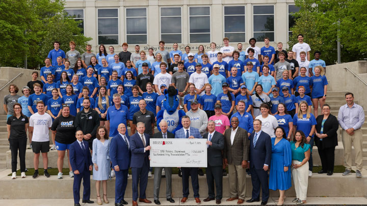 A large group of individuals in blue and gray attire poses for a photo, holding a check for $250,000 outside a building.