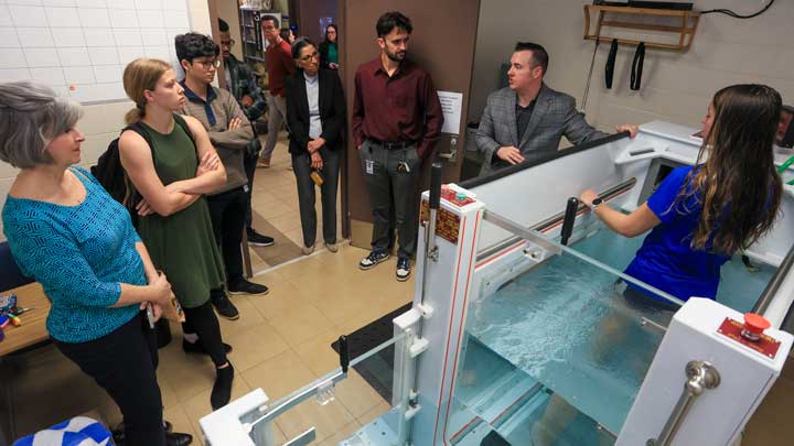 A group observes a demonstration by a woman in a blue shirt standing in a water tank, with medical equipment nearby.