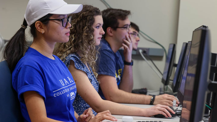 Three UAH students working on computers in a classroom, focused on their screens, wearing casual clothing.