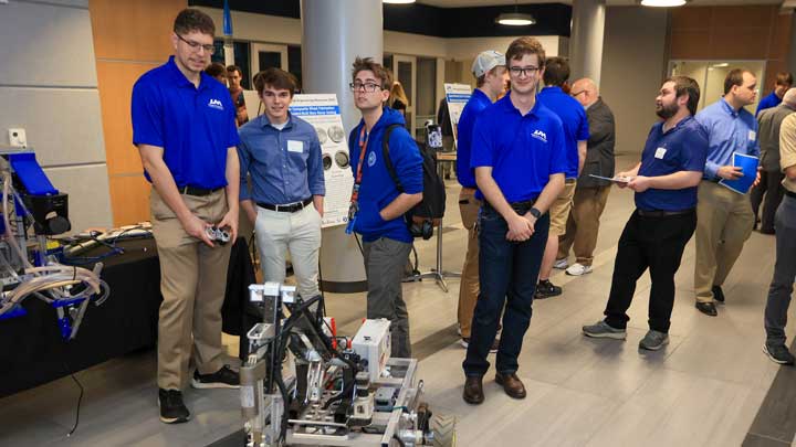 Several UAH students in blue shirts stand near a robotics display at an event, engaging with exhibits and each other.