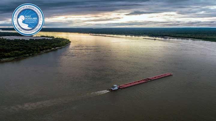 Aerial view of a river with a barge navigating through, surrounded by lush green banks under a cloudy sky. Gulf Scholars logo in corner.