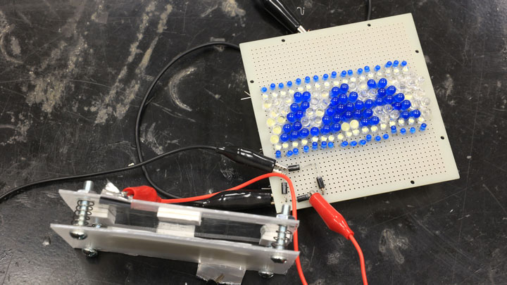 A circuit board displays UAH using colorful beads, with wires connected to a nearby component on a dark surface