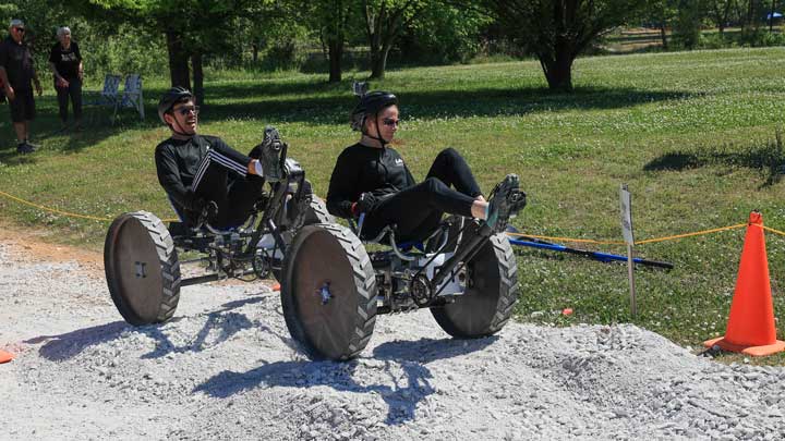 Two people wearing helmets ride large-wheeled pedal vehicles over a gravel path in a park with trees and grass.