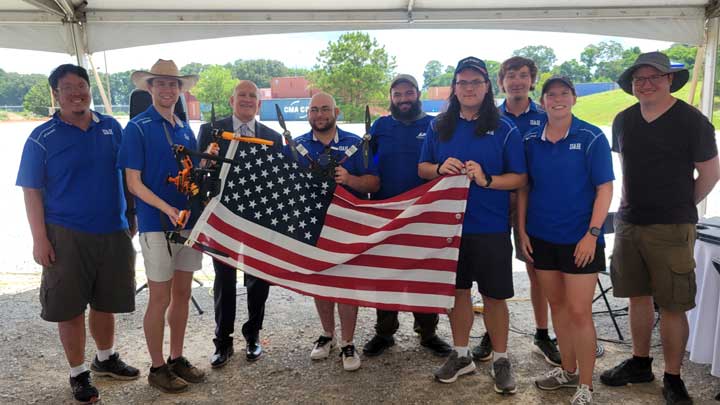 A diverse group of individuals in blue shirts poses under a tent, holding a large American flag and a drone.