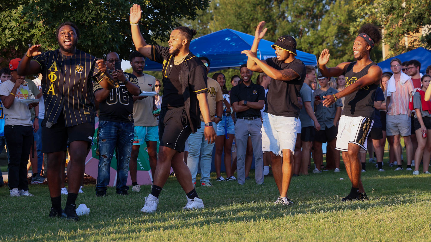 a group Alpha Phi Alpha members at U A H dance on a grassy lawn while smiling students watch.