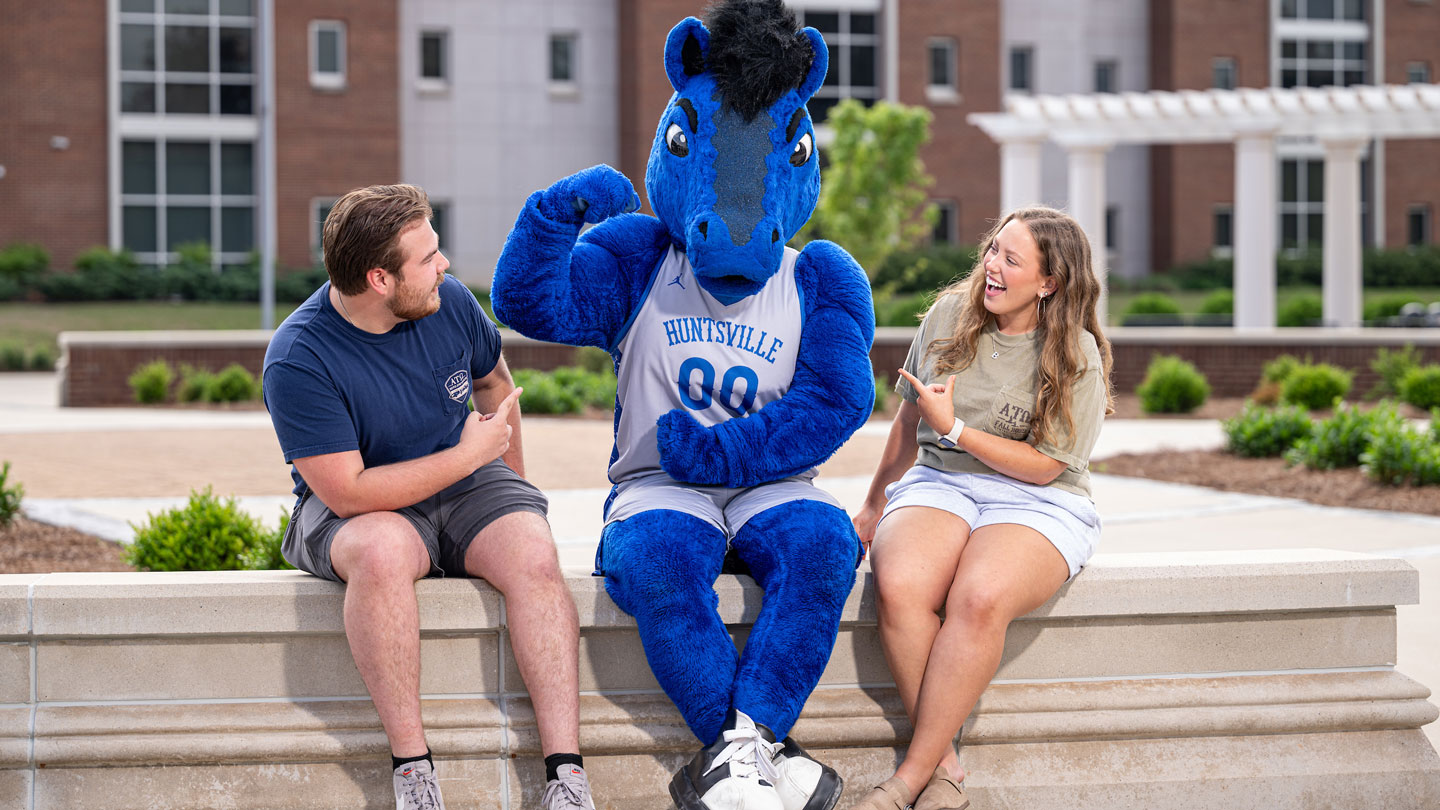 Charger Blue the U A H horse mascot flexes his muscles in front of two laughing students.