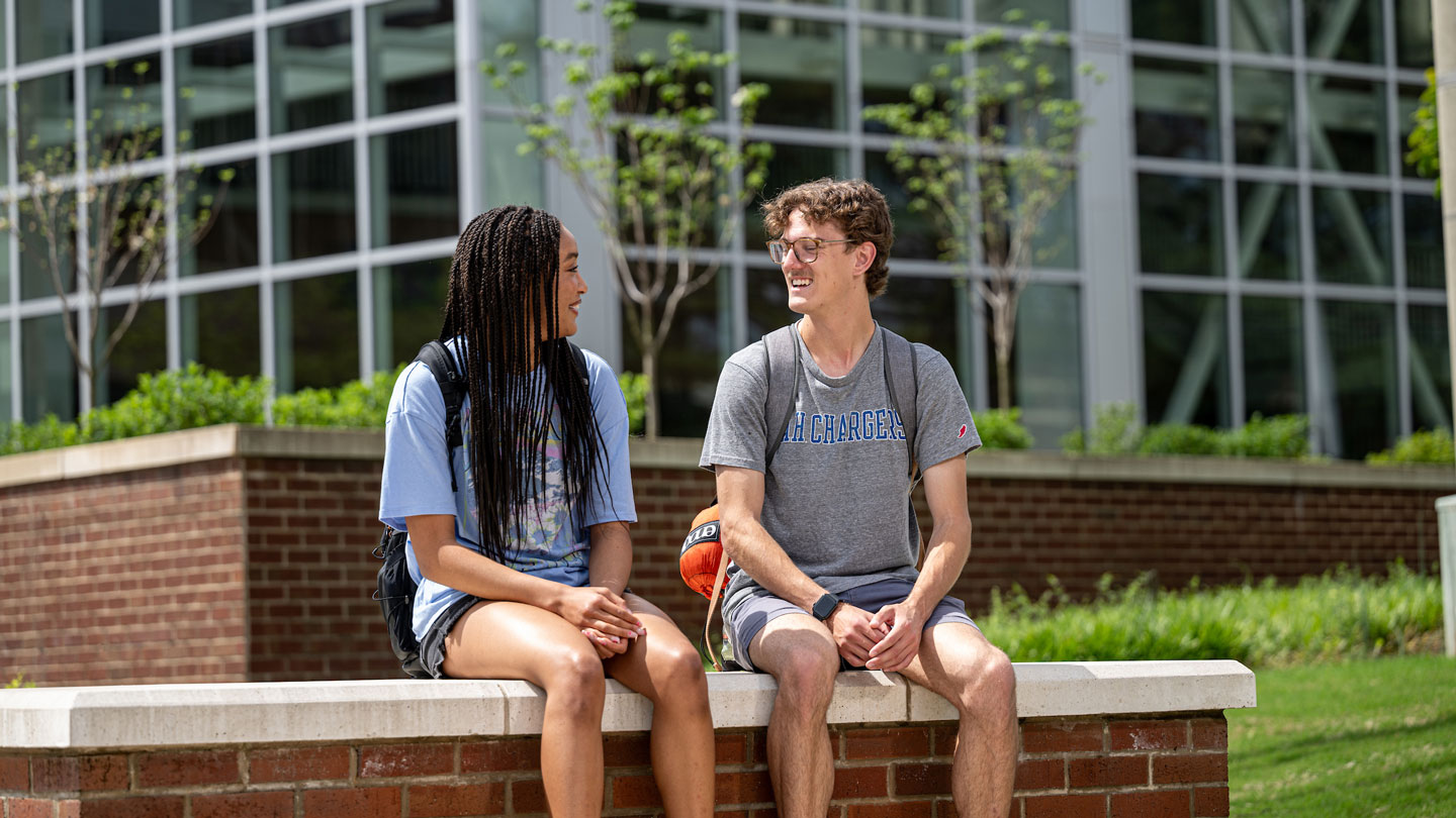 Two U A H students have a conversation while sitting on a low brick wall.