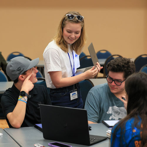 a smiling tutor holding a laptop talks to some students