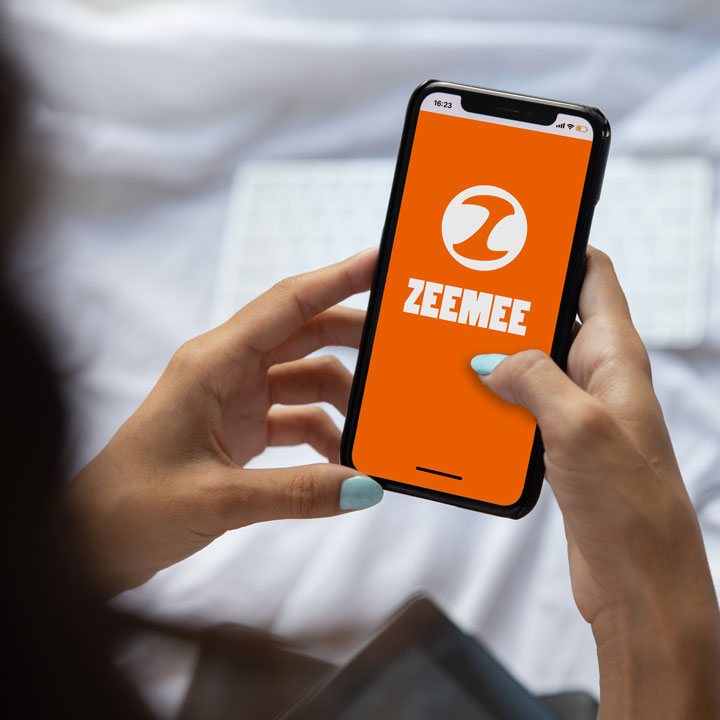 Hands hold a cell phone with the orange and white logo of the ZeeMee social media app on screen