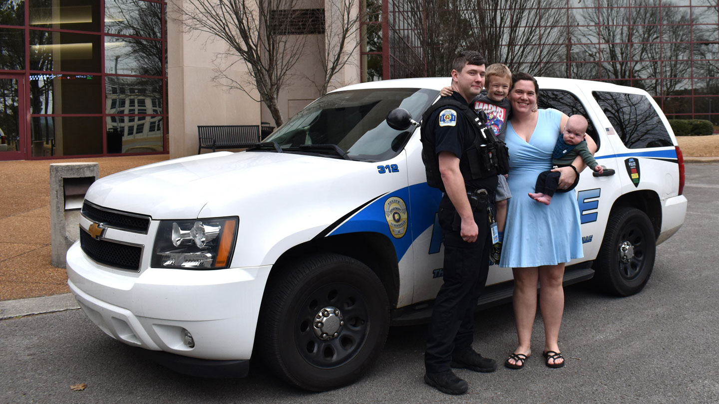 U A H ploice officer and business student Tyler Storm poses with his wife and two children in front of a U A H police vehicles parked in front of the U A H College of Business building.