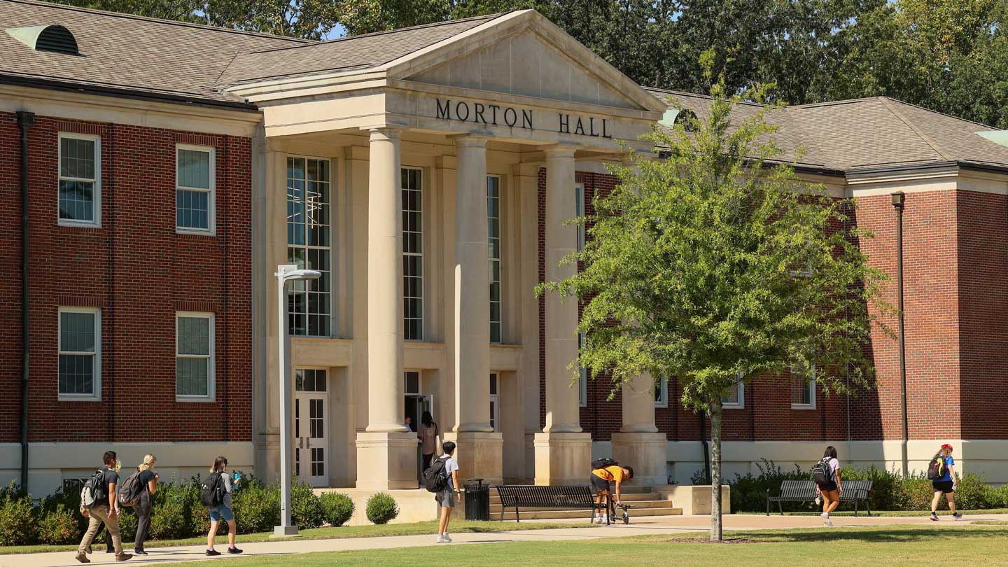 Students walking towards Morton Hall, a red brick building with tall columns, amidst a sunny campus setting.
