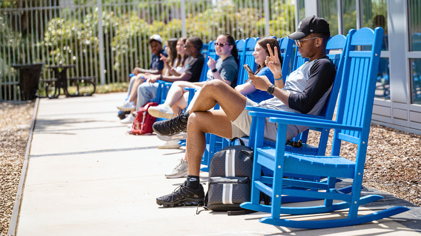 Students sitting outside in rocking chairs, rocking, while talking to each other and looking at their phones.