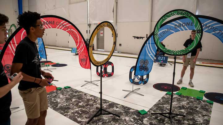 Two people operating drones in an indoor aerial drone competition course with colorful rings and timing screens.