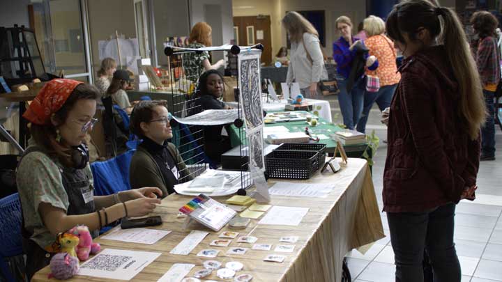 A ChargerCon attendee stops at a table and inspects artwork, art prints and buttons that two vendors are selling. In the background, other attendees browse numerous other similar tables.
