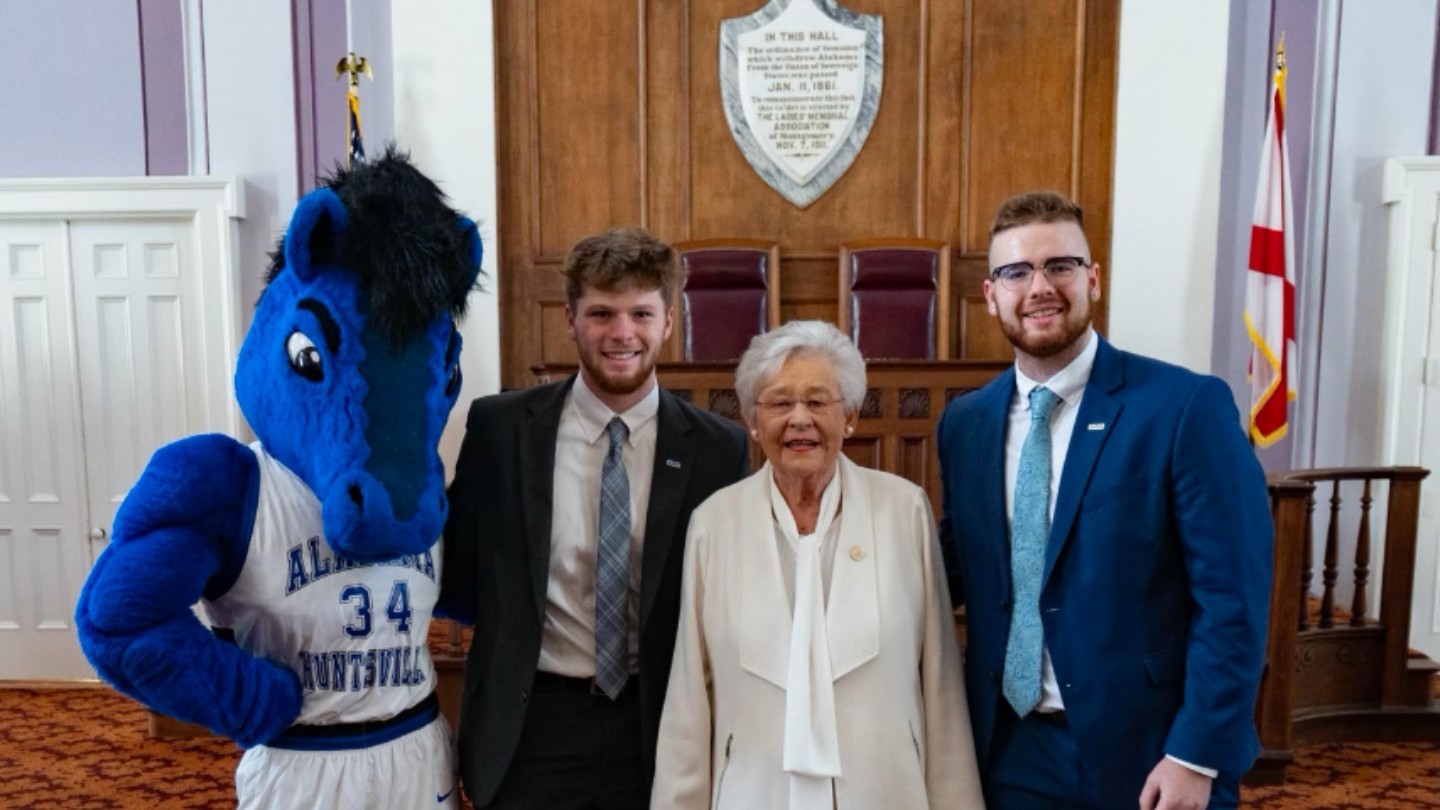 charger blue beside Kay Ivey and 2 students