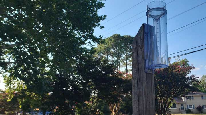 Clear rain gauge mounted on a wooden post with trees and residential houses in the background under a blue sky.