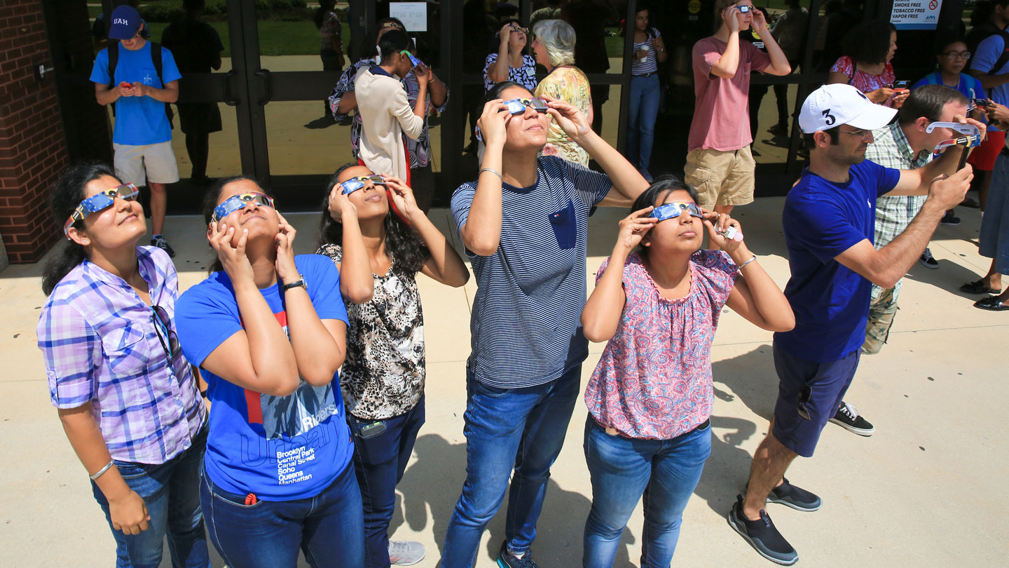 a groups of students wearing protective solar glasses look up at the sky to see a solar eclipse